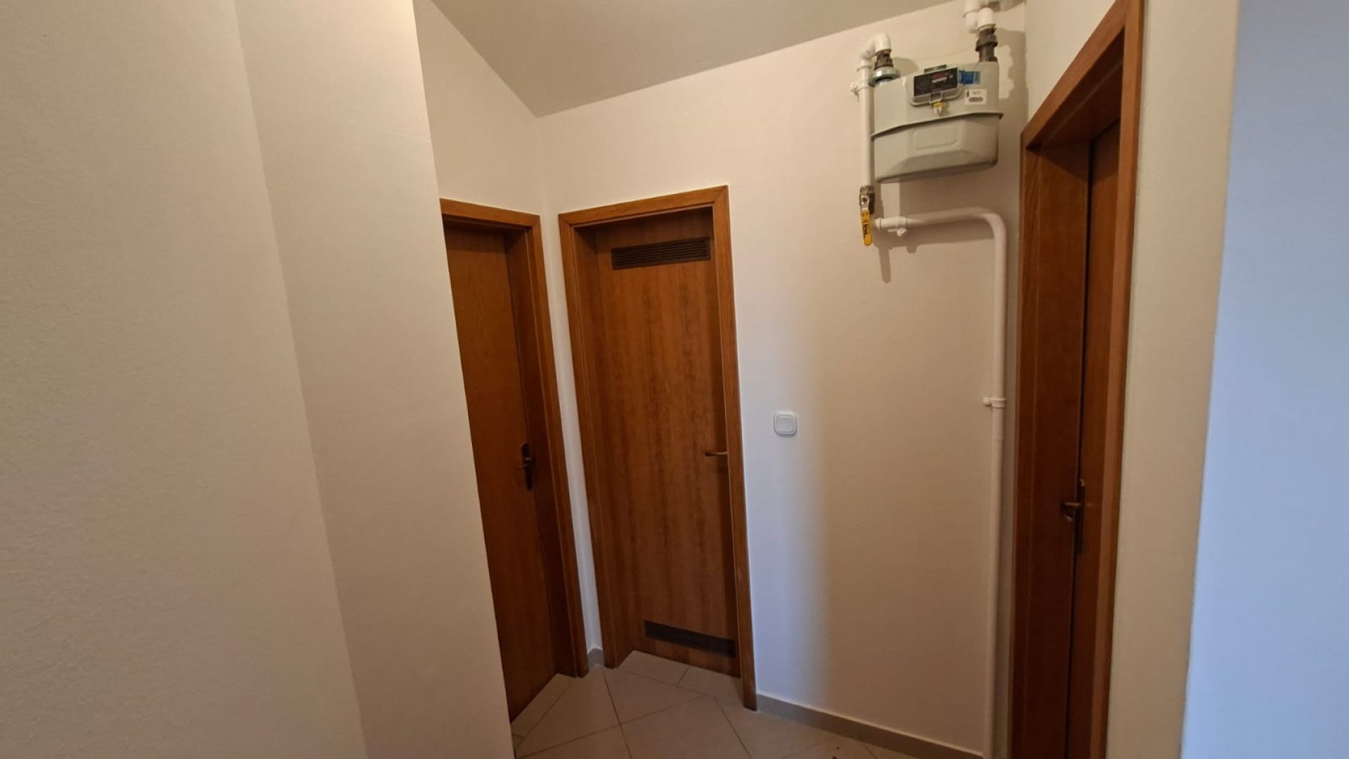 Hodnik s keramičkim pločicama, drvenim vratima i plinskim ormarićem na zidu.