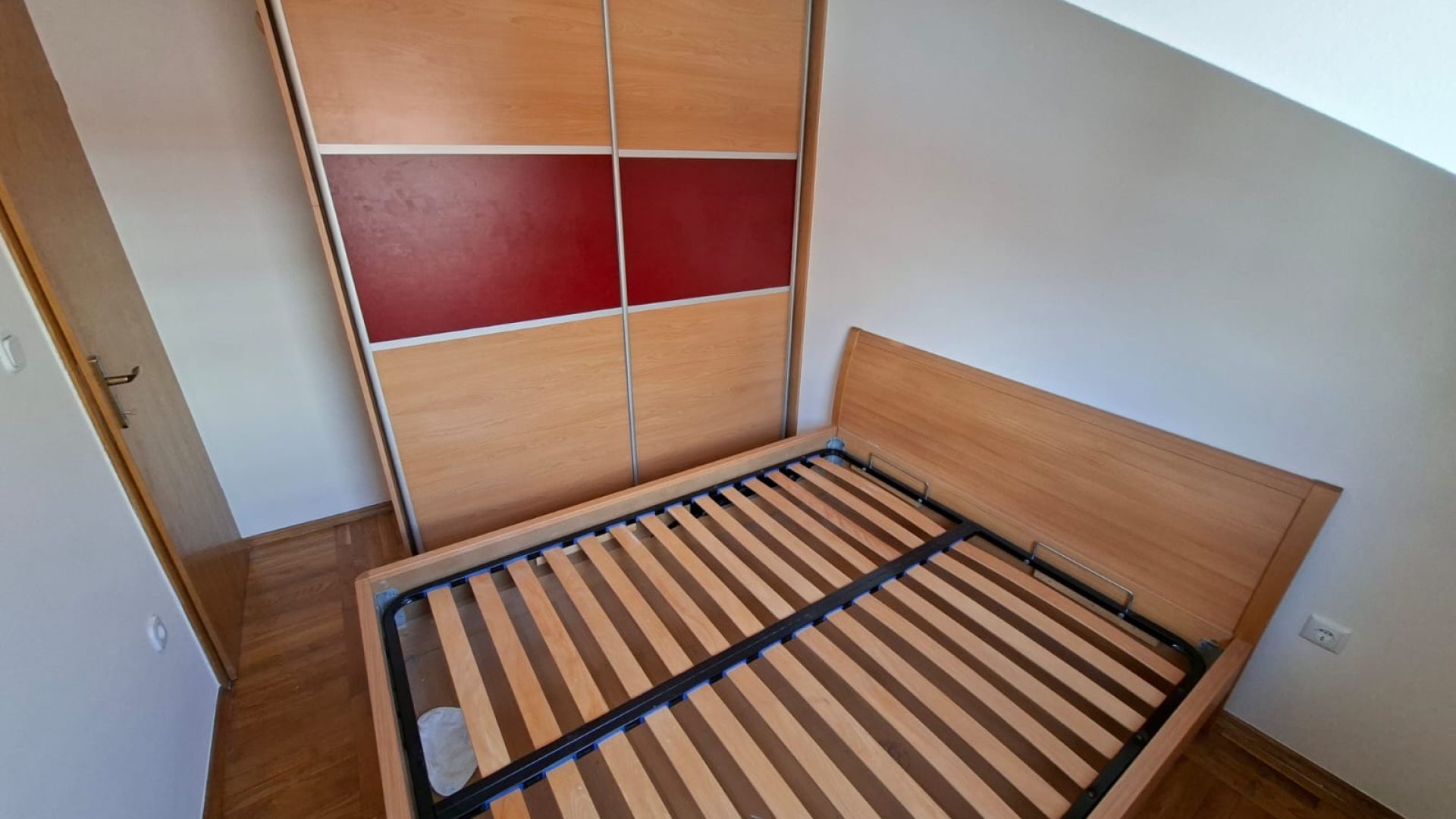 Spavaća soba s drvenim krevetom, kosim stropom i velikim ormarom s kliznim vratima.