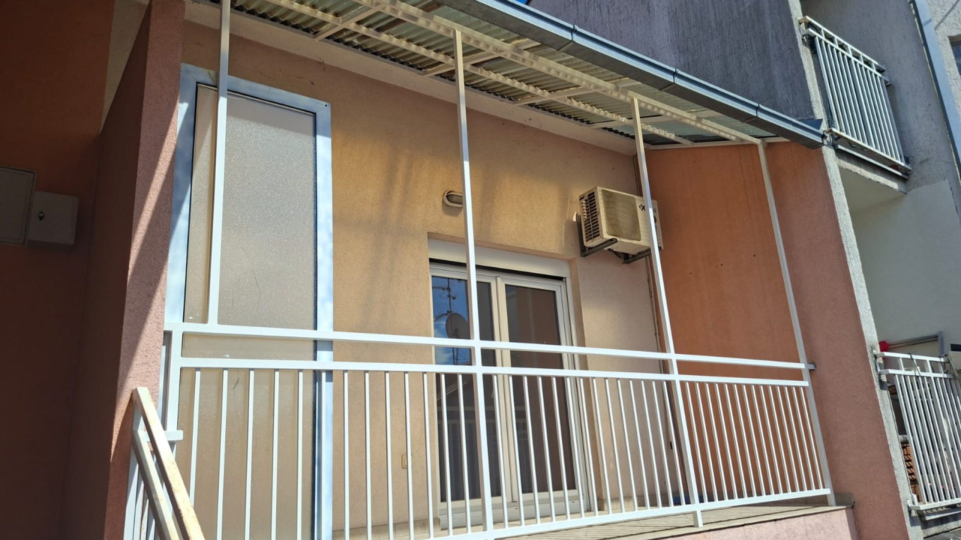 Balkon s bijelom metalnom ogradom i kliznim vratima prema dnevnom boravku.