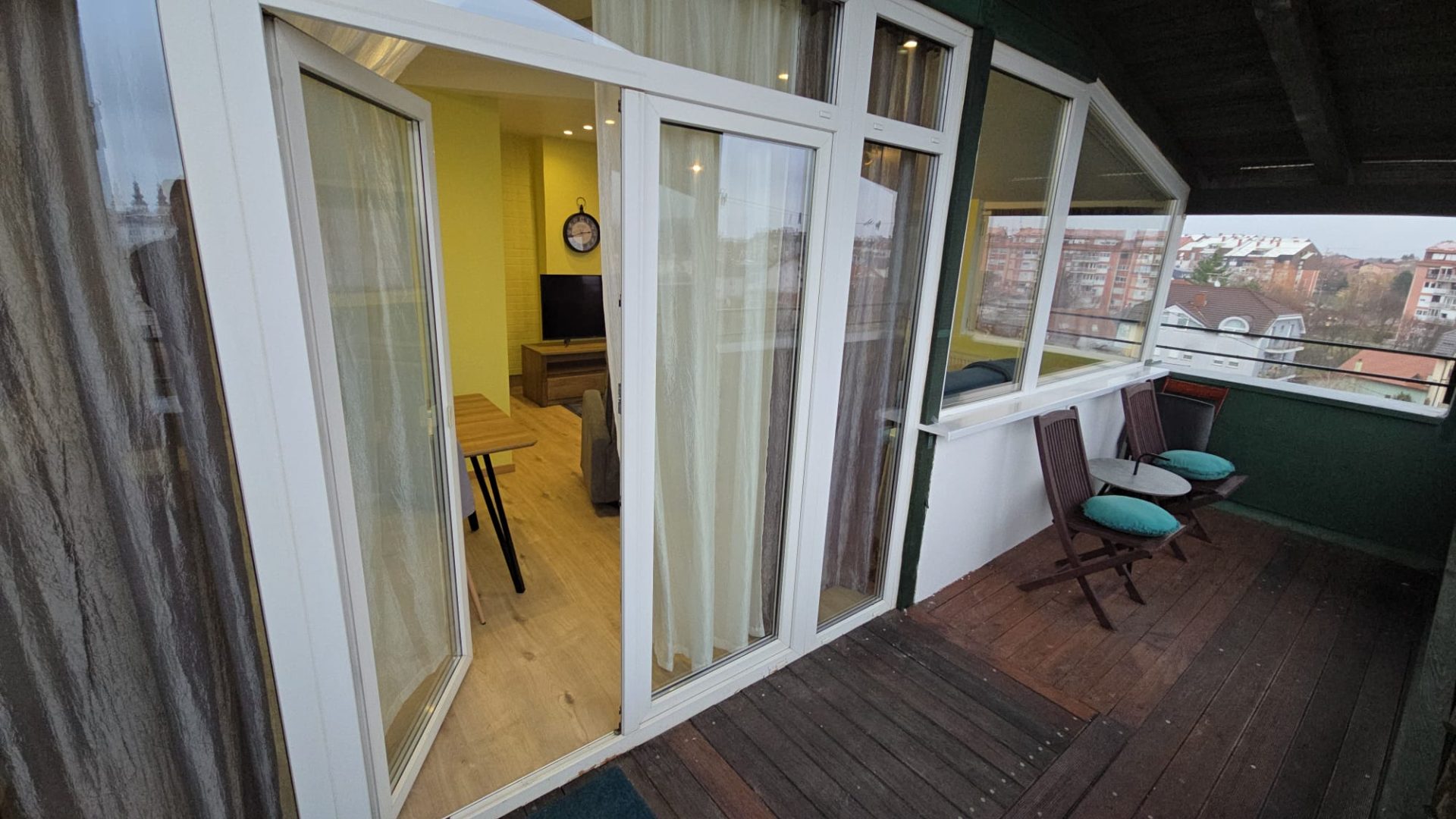 Balkon s drvenim podom, stolicama i pogledom u dnevni boravak kroz staklena vrata.