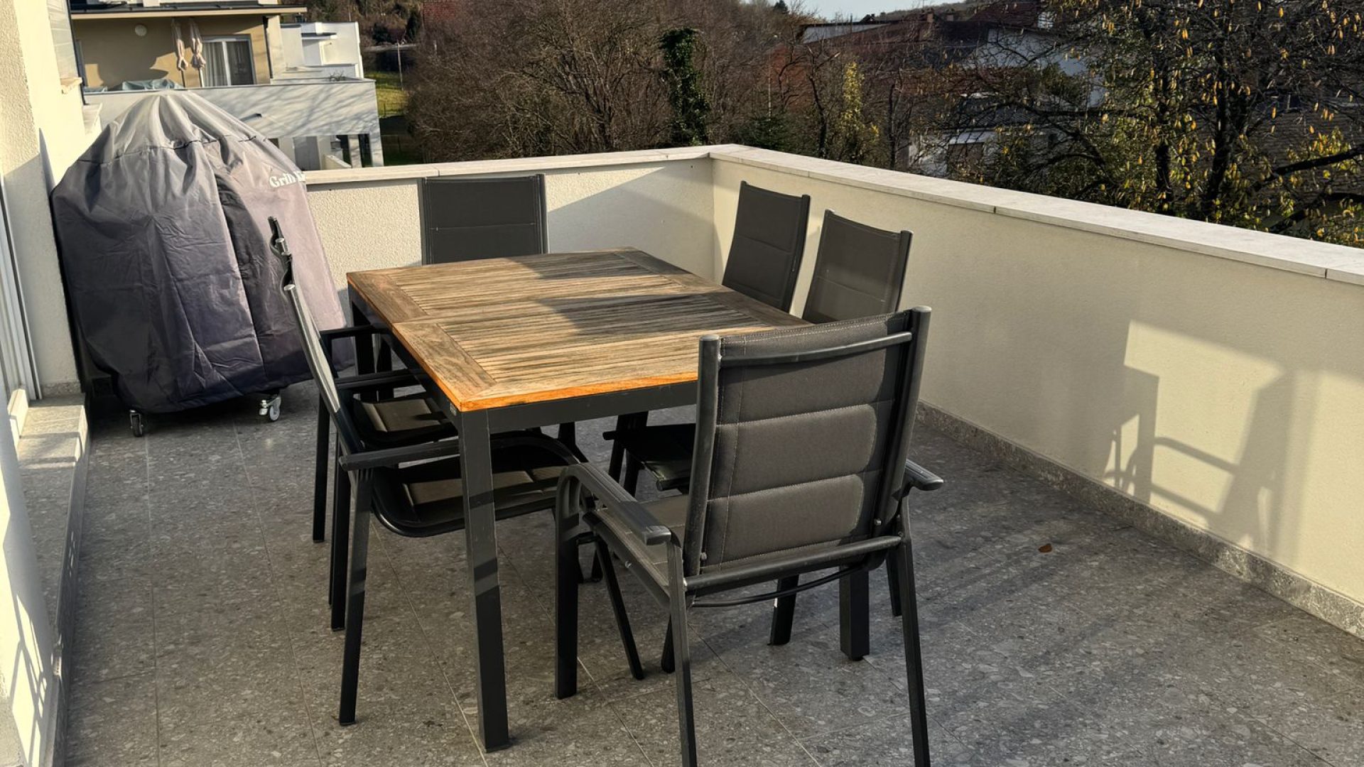 Prostrana terasa s blagovaonskim stolom i stolicama, pogledom na zelenilo i okolne kuće.